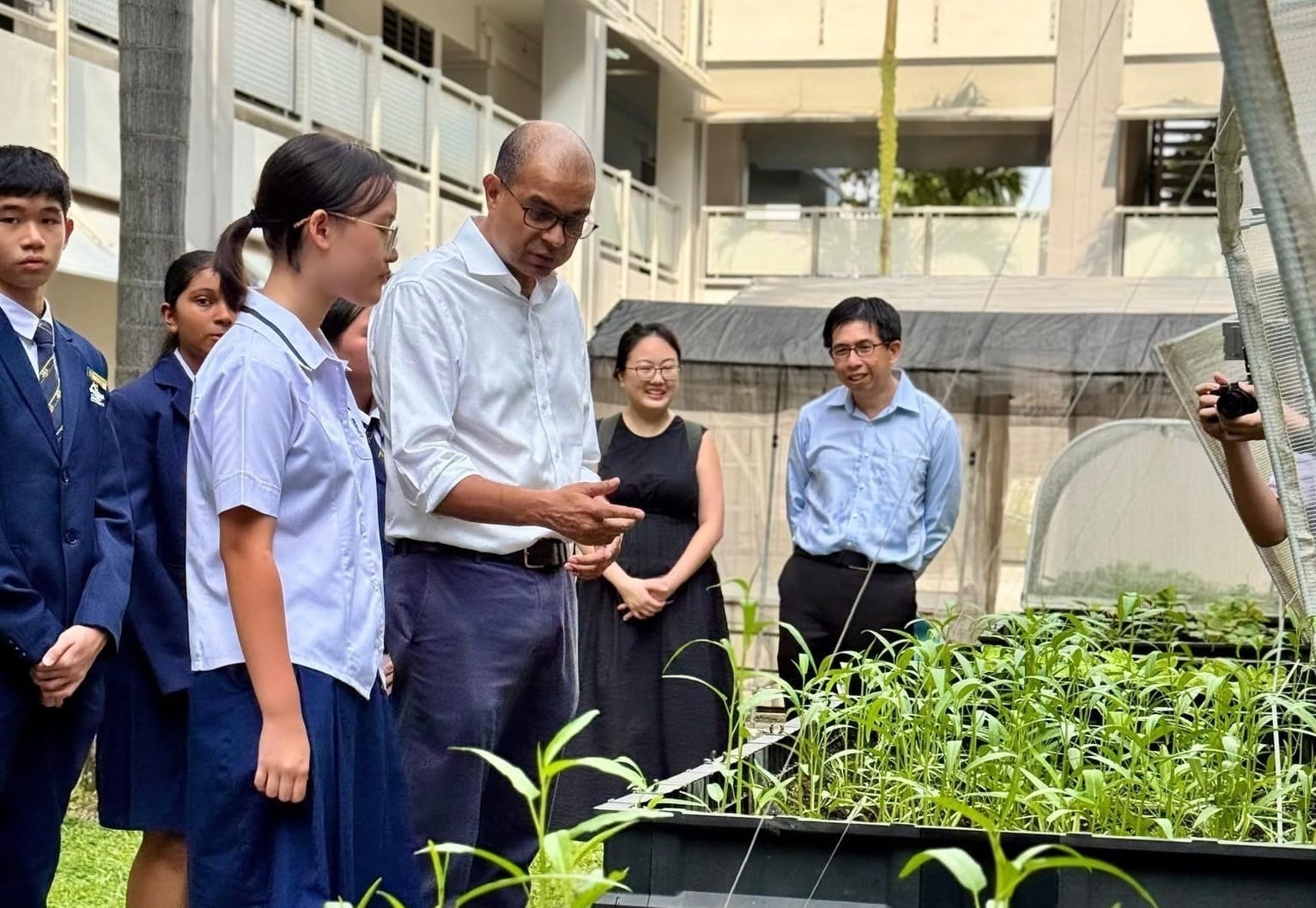 Photograph of a group of people, including students and adults, observing plants growing in raised garden beds within a courtyard. The setting highlights an educational or environmental activity focused on plant growth, with greenery and a multi-story building visible in the background.